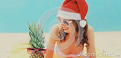 Christmas portrait of happy young smiling woman in red santa hat with funny pineapple lying on a beach together over sea Stock Photo