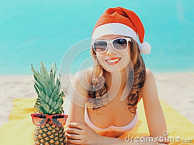 Christmas portrait of happy young smiling woman in red santa hat with funny pineapple lying on a beach together over sea Stock Photo