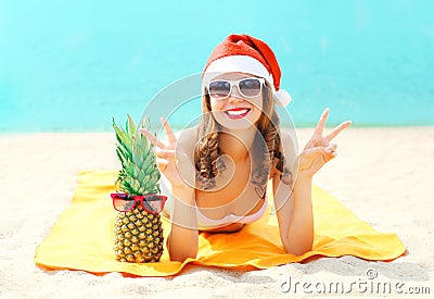 Christmas portrait pretty young smiling woman in red santa hat and pineapple lying on beach over sea Stock Photo