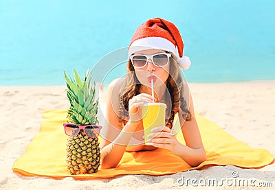 Christmas portrait pretty young woman in red santa hat with pineapple drinking from cup fresh fruit juice lying on beach over sea Stock Photo