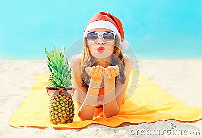 Christmas portrait pretty young woman in red santa hat with pineapple sends air kiss lying on beach over blue sea Stock Photo