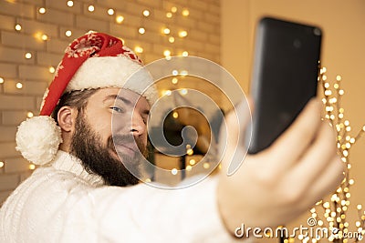 Christmas selfie by middle adult bearded man with white sweater and red hat in warm and cozy decorated room with yellow garland Stock Photo