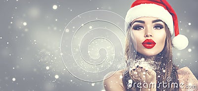 Beauty model woman in Santa`s hat blowing snow in her hand. Christmas winter fashion girl on holiday blurred winter background Stock Photo