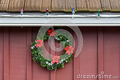 Christmas wreath made of artificial evergreen boughs and a light string, with red flowers and white snowflakes, on a red barn wall Stock Photo