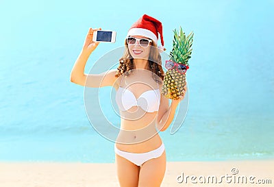 Christmas young smiling woman in red santa hat with pineapple taking picture self portrait on smartphone at beach over sea Stock Photo