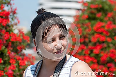The girl squints in the sun on the background of bright red flowers on the bushes Stock Photo