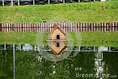 A city pond with a duck house, a steep bank with wooden piles and their beautiful reflections in the water on a clear sunny day. Stock Photo