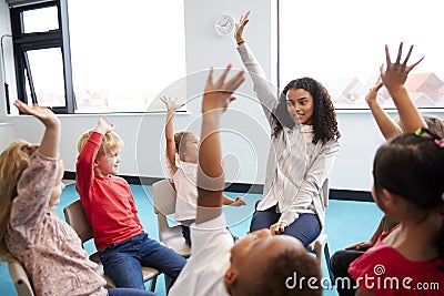 A class of infant school children sitting on chairs in a circle in the classroom, raising hands with their female teacher, close u Stock Photo