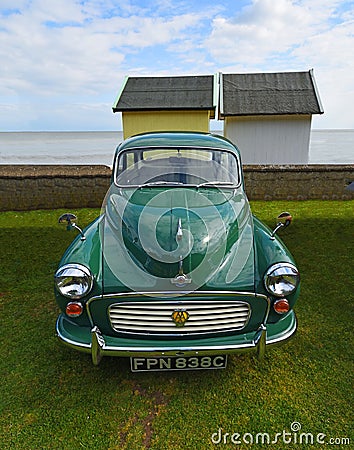 Classic Green Morris Minor with AA Badge parked with beach huts and ocean in background. Editorial Stock Photo