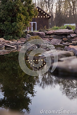 Clean pond in the garden of a country house. Stones and spruce on the lake. Quiet evening. The water reflects the sky Stock Photo