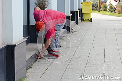 Cleaning of a city street - the worker the yard keeper in a uniform collects garbage by hands near a building. Editorial Stock Photo