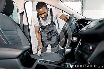 Cleaning of interior of the car with brush, car detailing service. Young pleasant black man doing car interior and seat Stock Photo