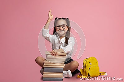 Clever pupil. Smiling girl in glasses with stack of books and backpack knows answer and raises hand up Stock Photo