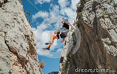 Climber teenager boy in protective helmet abseiling from vertical cliff rock wall using rope Belay device, climbing harness in Stock Photo