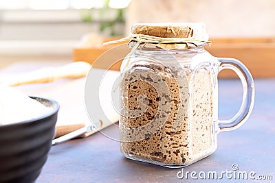Close-up of active sourdough starter in glass jar. Rye leaven for bread and kitchen utensil on rustic background. Selective focus Stock Photo