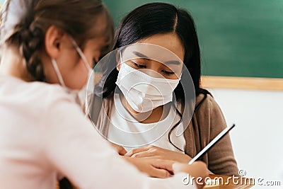 Close-up of Asian female teacher wearing a face mask in school building tutoring a primary student girl. Elementary Stock Photo