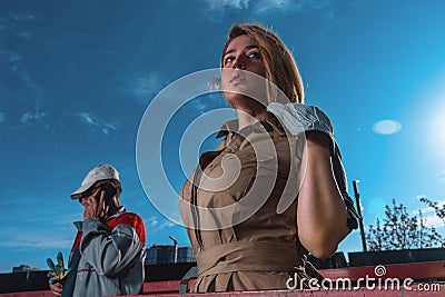 Close up Of Attractive Glamorous Model In Stylish Dress At Construction Cradle Near Worker Builder. Stock Photo