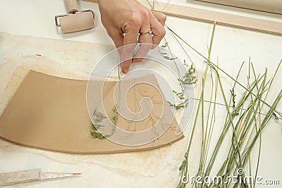 Close up ceramist hands imprinting herb on raw clay with rolling pin. Handmade, hobby art and handicraft concept Stock Photo
