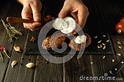 Close-up of a chef hands using a spoon adding sour cream to fried cutlets on a sorting board. The concept of preparing delicious Stock Photo