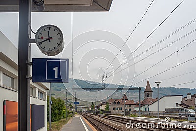 Close up of clock and platform sign in a small rural station on vintage houses, mountain and cloudy sky background Stock Photo