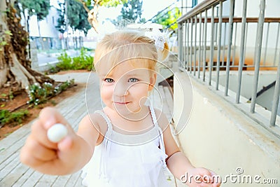 Close up cute little blondy toddler girl in white dress giving small sweet round candy for you. Selective focus. Copy space. Stock Photo