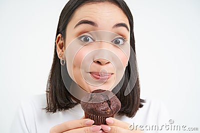 Close up of cute smiling asian woman, holding chocolate cupcake near mouth, having bite, enjoys eating pastry, white Stock Photo