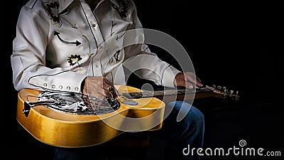 Close-up of a Dobro guitar resting on the lap of a musician dressed in cowboy attire on dark backdrop Stock Photo