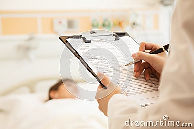 Close up of doctor writing on a medical chart with patient lying in a hospital bed in the background Stock Photo