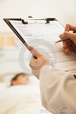 Close up of doctor writing on a medical chart with patient lying in a hospital bed in the background Stock Photo