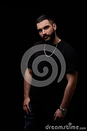 Close-up of a dramatic portrait of a young serious guy, musician, singer, rapper with a beard in black clothes on a black Stock Photo