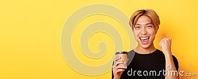 Close-up of energized handsome asian guy fist pump joyfully while drinking coffee, smiling excited over yellow Stock Photo