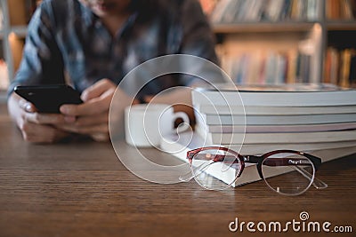 Close up eyeglasses and books stack on wooden desk in university or public library with man using mobile phone reading or text Stock Photo