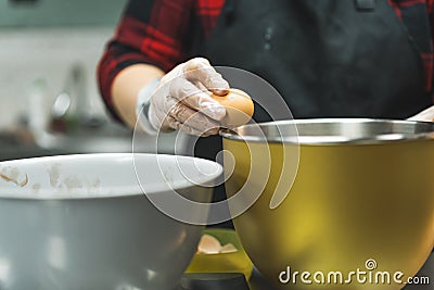 Close-up of female baker wearing gloves and black apron cracking egg into golden bowl to prepare cupcake batter Stock Photo