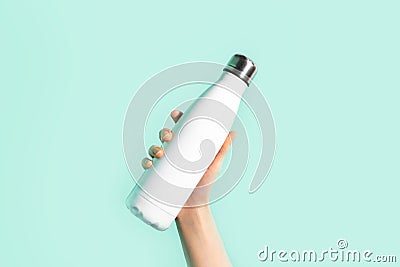 Close-up of female hand, holding white reusable steel stainless eco thermo water bottle with mockup, isolated on cyan color. Stock Photo