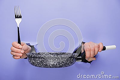 Close up of female hands holding Frying pan and fork through torn paper purple background. Kitchen utensils concept. Stock Photo