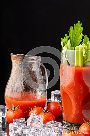 Close-up of glass with Bloody Mary cocktail with celery, jug with tomato juice, ice and cherry tomatoes, on black background, in v Stock Photo