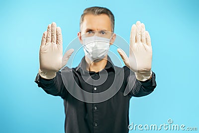 Close-up of the gloved hands of a young man in a medical protective mask on a blue background. Social distance. Stop gesture Stock Photo