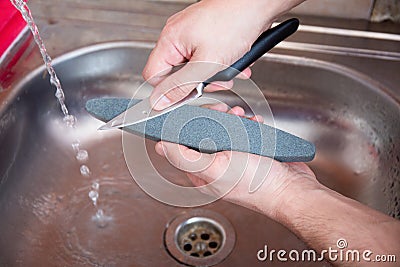 Close-up hands of a man carefully sharpen a knife under a stream of water on a grindstone. Home household sharpening Stock Photo