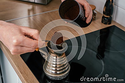 Close up of hands of unrecognizable man putting full heaped tea spoon of ground coffee from round tin in copper cezve. Stock Photo