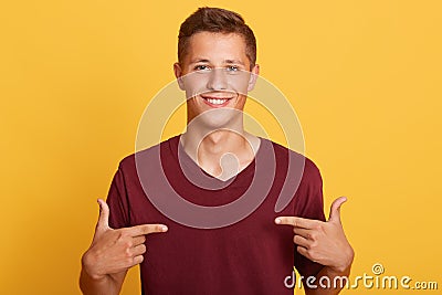 Close up of handsome man in blank burgundy t hirt pointing at himself isolated over yellow studio background, male with pleasant Stock Photo