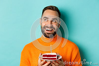 Close-up of happy adult man celebrating birthday, holding bday cake with candle and making wish, standing against Stock Photo