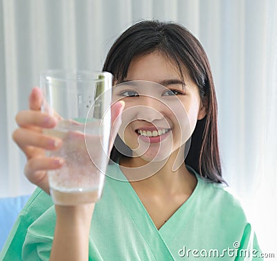 Close up of happy young inpatient woman who is getting well from her ailment smiling on to camera and holding a glass of water in Stock Photo