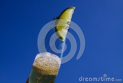 Close up of isolated bottleneck with sparkling yellow beer and a slice of lime against cloudless deep blue sky Stock Photo