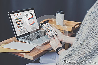 Close-up of laptop on wooden table and smartphone with graphs, diagrams and charts on screen in hands of businesswoman. Stock Photo