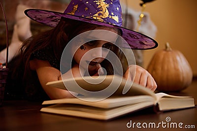 Close-up little witch, enchantress in wizard hat reading sorcery and spell magic book surrounded by Halloween atmosphere Stock Photo