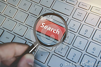 Close up of male hand holding magnifying glass above the laptop red key Search. Concept of internet searching. Enlarging Stock Photo