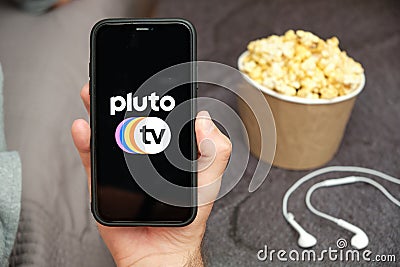 Close up mans hand holding a mobile phone with Pluto tv logo with Apple earphones and popcorn box next to him, Free TV Editorial Stock Photo