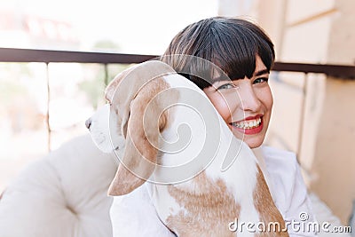 Close-up outdoor portrait of smiling girl with dark-brown hair holding beagle dog. Stunning brunette lady in white Stock Photo