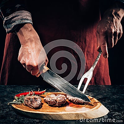 Chef in apron with meat fork and knife slicing gourmet grilled steaks with rosemary and chili pepper on wooden board Stock Photo