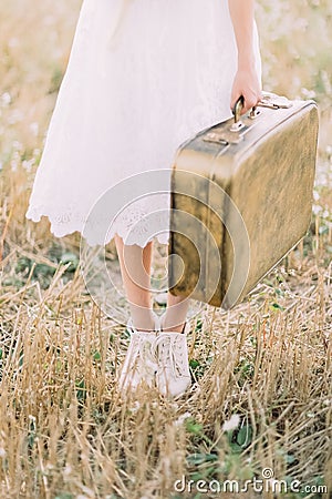 The close-up photo of the legs of the bride dressed in the knee-length dress and carrying the vintage suitcase in the Stock Photo